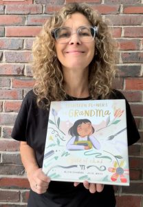 Author Alison Goldberg standing outside by a brick wall holding picture book, Eighteen Flowers for Grandma.